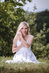 young blond girl sitting on the grass and smiling, a fashion model, a woman in a white dress