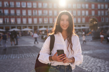 Young beautiful woman in the city of Madrid looking at camera and holding smartphone.