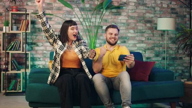 In the living room excited couple watch football match on the smartphone they are happy when their team wining and get the victory.