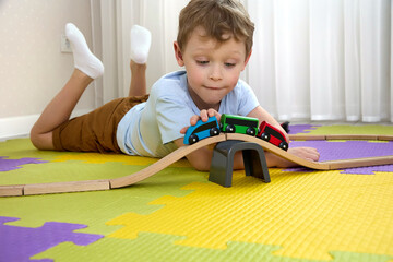 Close-up of wooden toys on puzzle mats that the boy is playing with. A child lying on the floor rolls a train with colorful cars along a wooden railway.