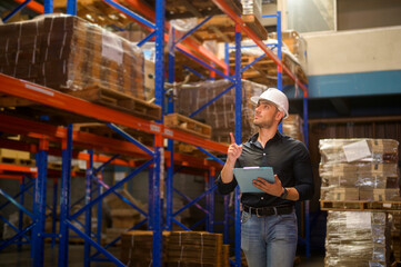 Young worker wearing helmet checking inventory and counting product on shelf in modern warehouse.
