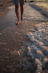 Legs of a man in shorts and walking along the beach near the waves of water