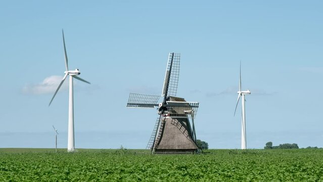 Two modern white windturbines behind an old traditional dutch windmill in an agricultural field under a blue sky in summer. Old and new or contrast concept. 