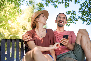 Cheerful young couple smiling and laughing using smart phone sitting in a bench in a park. Man and...