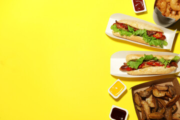 Tasty hot dogs, potato wedges, fried onion rings and different sauces on yellow background, flat lay with space for text. Fast food