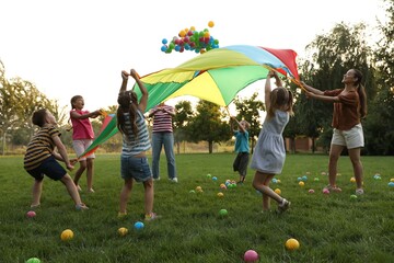 Group of children and teacher playing with rainbow playground parachute on green grass. Summer camp activity