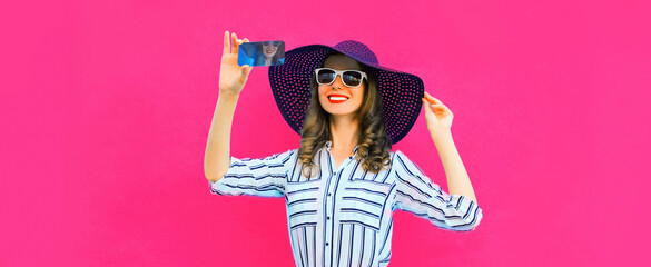 Portrait of beautiful happy smiling woman taking selfie with smartphone wearing black round straw summer hat, white striped shirt on pink background