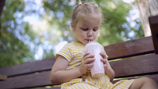 Little girl drinks a milkshake through a straw. Close-up portrait of cute child girl sitting on park bench and drinking milkshake
