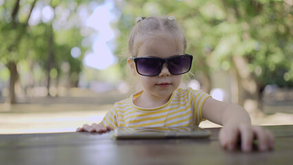 Little girl in mom's sunglasses is learning foreign language by repeating words from mobile phone. Close-up portrait of child girl sitting in city park and learning foreign language using cellphone