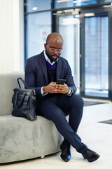 African businessman typing a message on his mobile phone while sitting on the couch in the lobby and waiting for the meeting