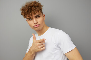 a self-confident, curly-haired young man, standing on a gray isolated background and smiling pleasantly looks away, pointing with his index finger. Close portrait