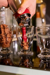 A portrait of someone spraying chocolate mousse into glass cups using an espuma device to make it more airy. The tasty dessert is brown, delicious and ready to eat.