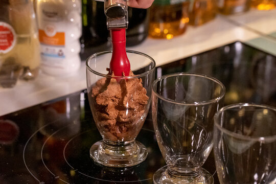 A close up portrait of someone spraying chocolate mousse into glass cups using an espuma device to make it more airy. The dessert is brown, delicious and ready to eat.