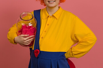 funny clown in a wig and a yellow-blue suit with a propeller on his back holds a jar of jam in his...