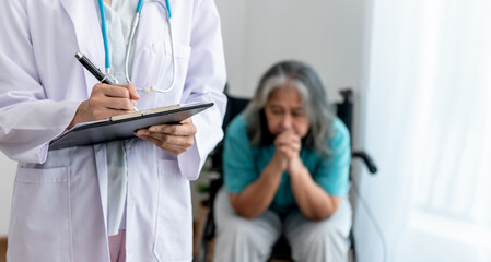 The doctor writing report for record the patient's condition, with blur background of Asian female elderly patient sitting in a wheelchair and have stress, to elderly and health care concept.