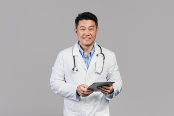 Cheerful middle aged asian male doctor in white coat with stethoscope, tablet isolated on gray background