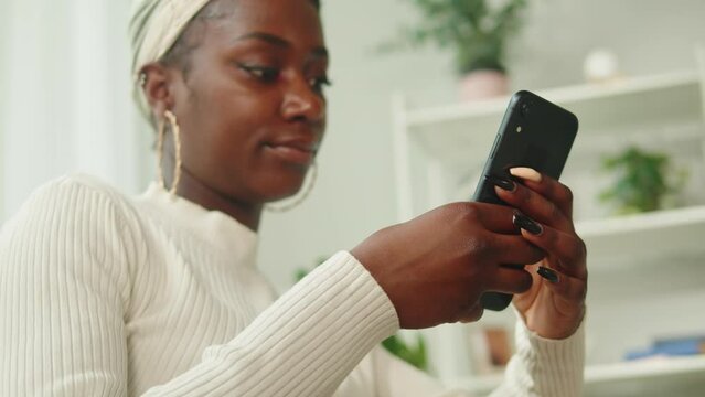 Happy native African American woman using phone close-up. Young female student chatting online at smartphone and smiling, sitting on sofa in living room. Communicating with friends via internet. 