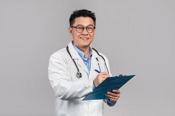 Cheerful mature korean man doctor in white coat, glasses with stethoscope writes at tablet