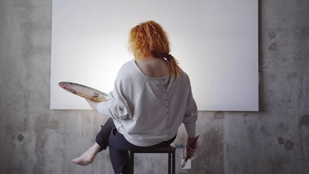 Female painter sitting in front of white canvas holding paintbrushes and palette. Caucasian ginger hair artist waiting for inspiration. High quality 4k footage