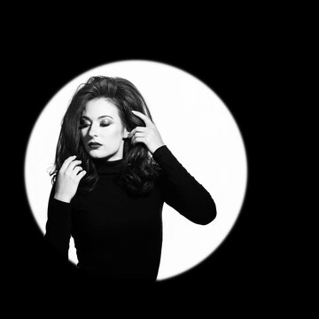 Fashion, make-up and entertainment concept. Beautiful woman portrait standing in illuminated circle in black background. Model with evening makeup touching her volume wavy hair. Black and white image
