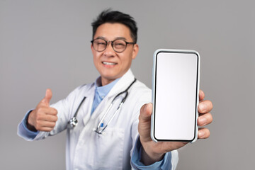Fototapeta na wymiar Glad confident mature asian male doctor in white coat, glasses show phone with empty screen, thumb up