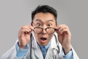 Portrait of shocked excited funny adult asian man doctor in white coat with open mouth, takes off glasses