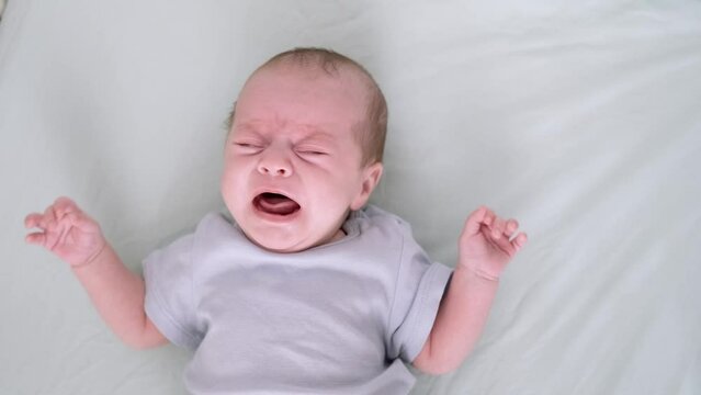 Dissatisfied, crying, upset, sad Newborn baby boy lying in nursery in crib with blue clothes on bed in bedroom at home, child crying is hungry or want to sleep