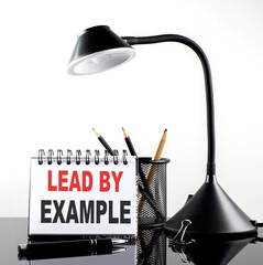 LEAD BY EXAMPLE text on notebook with pen and table lamp on the black background