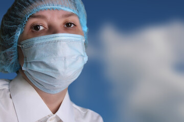 Close-up portrait of a woman doctor in a medical cap and mask against the blue sky.