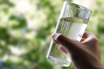 A glass of water in a hand close-up on a natural green background outdoors.