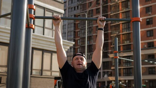 A middle-aged man in a baseball cap pulls himself up on a horizontal bar in the courtyard of the neighborhood.