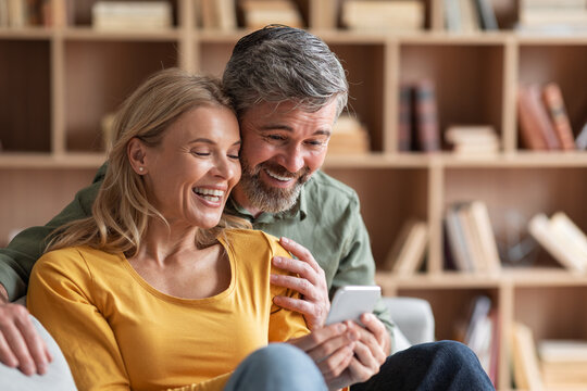 Happy Middle Aged Couple Watching Family Photos On Smartphone At Home