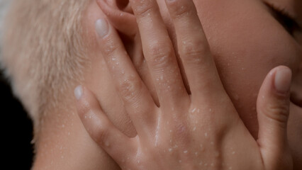 Macro shot of good-looking young slim blond Caucasian woman touches her wet neck, face and ear on black background | Wet skin texture shot for moisturizer commercial