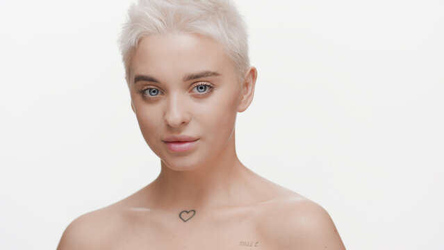 Gorgeous slim young light-haired Caucasian woman with bare shoulders looks at the camera smiling on white background | Skin care concept
