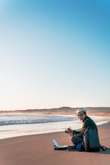 Fototapeta premium digital nomad sitting on the beach with his laptop and taking notes diary on the shore of the beach at sunset