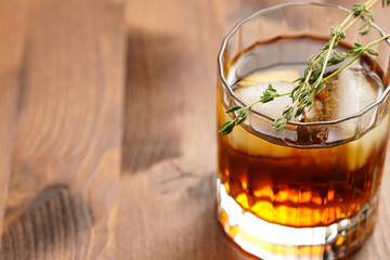 a tumbler glass with brown alcoholic drink, thyme and ice cubes - whisky, rum or cognac - on wooden table, rural scene