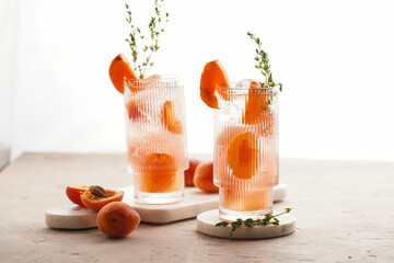 Two glasses with refreshing summer cocktail with ice cubes, apricot slices and fresh thyme on marble board on pale purple background