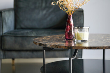 Mock up in interior: blank off-white pillar candle in glass jar with label and silver colored lid on a marble table with red vase in front of couch - a living room set-up
