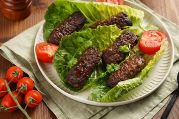 Traditional south european skinless sausages cevapcici made of ground meat and spices on lettuce leaves with thyme and tomatoes on white plate on kitchen towel, black silverware