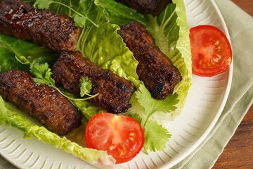 Traditional south european skinless sausages cevapcici made of ground meat and spices on lettuce leaves with thyme and tomatoes on white plate on kitchen towel, black silverware