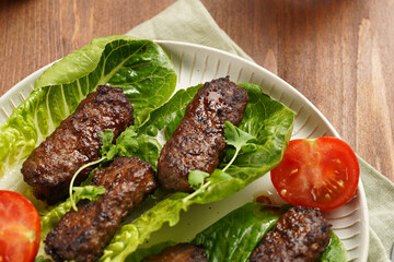 Traditional south european skinless sausages cevapcici made of ground meat and spices on lettuce leaves with thyme and tomatoes on white plate on kitchen towel, black silverware