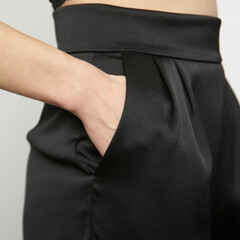 Close up image of female black satin trousers with wide waistband and hand in pocket.