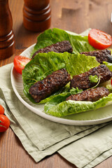 Traditional south european skinless sausages cevapcici made of ground meat and spices on lettuce leaves with thyme and tomatoes on white plate on kitchen towel, black silverware