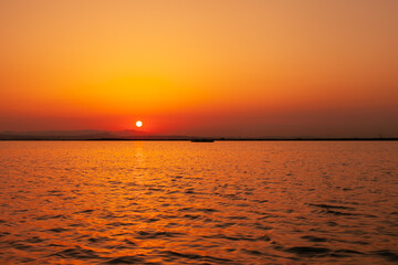 Colorful orange, yellow and red sunset over the water of lake with sun setting in the water. Natural park at sunset with distant silhouettes of mountains. Sunset landscape view from a boat. Traveling 
