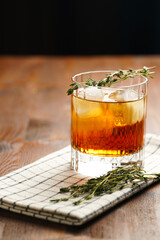 a tumbler glass with brown alcoholic drink, thyme and ice cubes on linen napkin - whisky, rum or cognac - on wooden table, rural scene