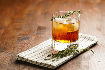 a tumbler glass with brown alcoholic drink, thyme and ice cubes on linen napkin - whisky, rum or...