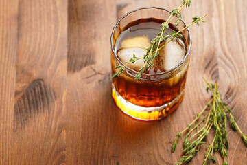 a tumbler glass with brown alcoholic drink, thyme and ice cubes - whisky, rum or cognac - on wooden table, rural scene