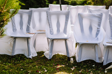 Many wooden chairs in white fabric stand at the wedding ceremony on the grass with rose petals.