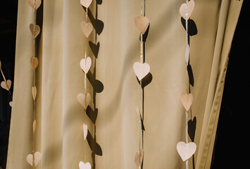 Lots of beautiful white paper hearts hanging from a ribbon on a fabric background with sunlight and shadow. Festive photo.