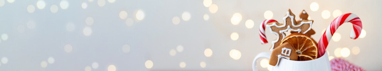 Hot winter drink in a white mug: cozy home composition with homemade gingerbread cookies, candy cane, fir tree branch. Wooden background, Christmas lights and candles. Banner copy space